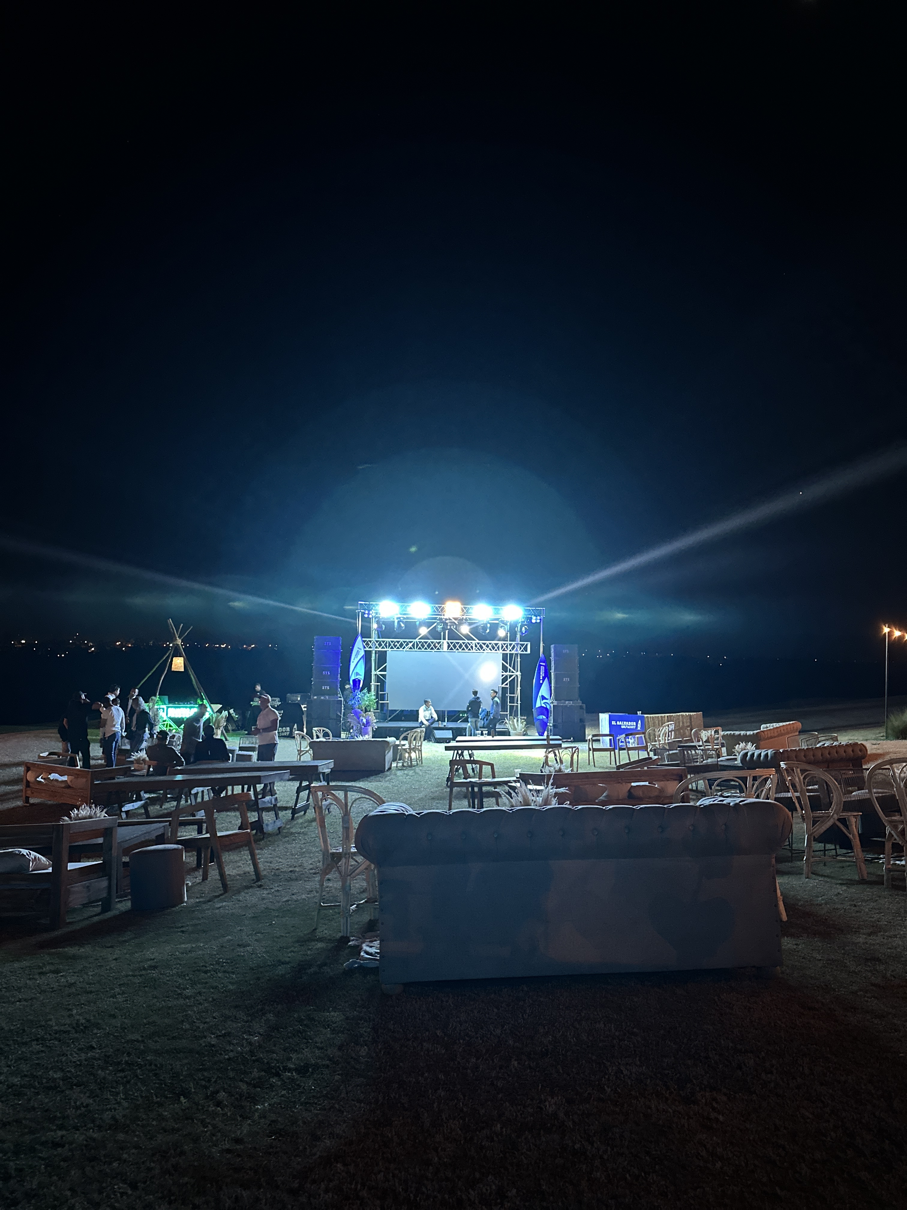 Evento al aire libre con escenario y sillas, luces de contra y cielo nocturno